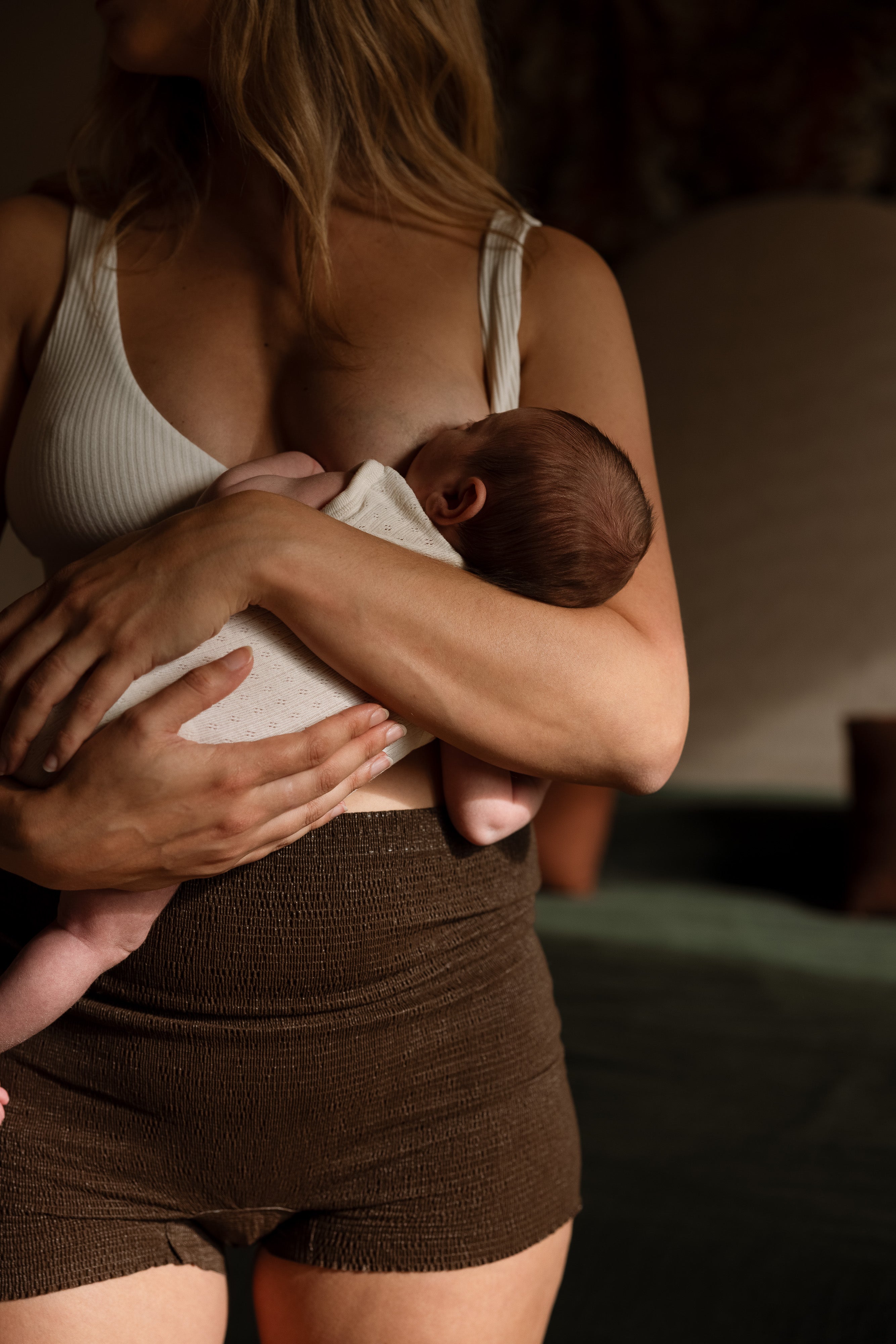 A woman wearing a white ribbed tank top and brown ribbed shorts holds a swaddled newborn baby against her chest in a dimly lit indoor setting.